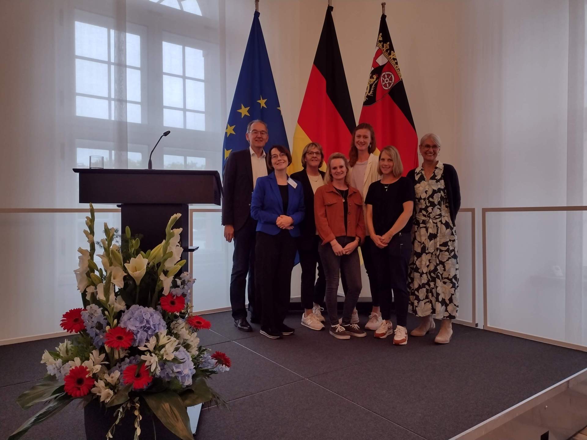Gruppenfoto bei einem Festakt in der Staatskanzlei in Mainz, aufgenommen vor den Flaggen der Europäischen Union, Deutschlands und Rheinland-Pfalz.