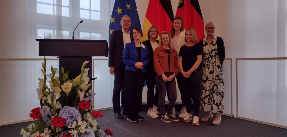 Von links: Erster Beigeordneter des Kreises Südliche Weinstraße Georg Kern, Heike Neumann, Leiterin des Referats Demografie bei der Kreisverwaltung SÜW, Fachkräfte Gemeindeschwester plus Ute Wingerter, Marleen Kunstätter, Jana Hoyer, Katharina Rösch und Claudia Sarter beim Festakt des rheinland-pfälzischen Ministeriums für Arbeit, Soziales, Transformation und Digitalisierung anlässlich des zehnjährigen Bestehens der Gemeindeschwester plus. Nicht auf dem Bild ist die Fachkraft Nadja Hieb. Foto: KV SÜW Gruppenfoto bei einem Festakt in der Staatskanzlei in Mainz, aufgenommen vor den Flaggen der Europäischen Union, Deutschlands und Rheinland-Pfalz.