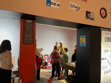 Blick in einen Raum, in dem Kinder um einen Mann mit Warnweste und eine Frau stehen. An der Wand hängen Kennzeichenschilder und Schriftzüge aus verschiedenen europäischen Städten, im Hintergrund ein Wandbild mit verschiedenen Nationalflaggen.