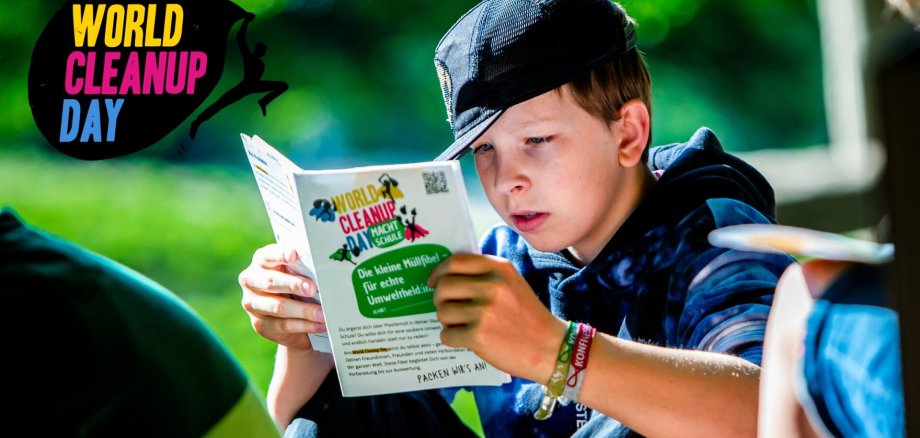 Ein Junge sitzt im Freien, trägt eine schwarze Kappe und liest konzentriert eine Broschüre zum World Cleanup Day. Auf der Broschüre steht unter anderem "Die kleine Müllfibel – für echte Umweltheld*innen". Links oben im Bild ist das bunte Logo des World Cleanup Day zu sehen.