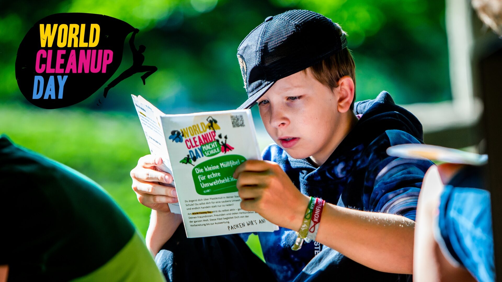 Ein Junge sitzt im Freien, trägt eine schwarze Kappe und liest konzentriert eine Broschüre zum World Cleanup Day. Auf der Broschüre steht unter anderem "Die kleine Müllfibel – für echte Umweltheld*innen". Links oben im Bild ist das bunte Logo des World Cleanup Day zu sehen.