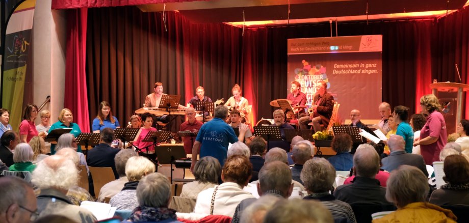 Blick aus dem Publikum auf eine Bühne, auf der mehrere Musikerinnen und Musiker musizieren. Vor der Bühne singt ein gemischter Chor unter Leitung eines Dirigenten in blauem Shirt, der mit dem Rücken zum Publikum steht. Auf einer Leinwand auf der Bühne ist das Logo zu "Deutschland singt und klingt" zu sehen. Außerdem die Aufschrift "Gemeinsam in ganz Deutschland singen"