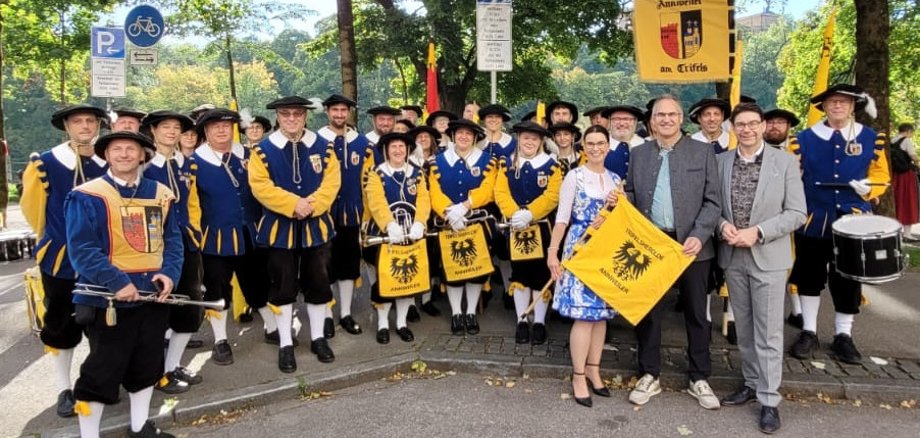 Eine große Gruppe von Musikerinnen und Musikern in blauen und gelben Kostümen posiert gemeinsam im Freien. Vorne stehen mehrere Personen mit einem gelben Banner, auf dem „Annweiler am Trifels“ zu lesen ist.