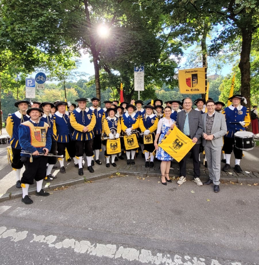 Eine große Gruppe von Musikerinnen und Musikern in blauen und gelben Kostümen posiert gemeinsam im Freien. Vorne stehen mehrere Personen mit einem gelben Banner, auf dem „Annweiler am Trifels“ zu lesen ist.