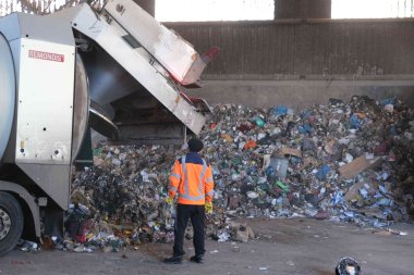 Ein Mann in orangener Jacke steht in einer Halle mit gelagertem Restmüll neben einem Müllfahrzeug.