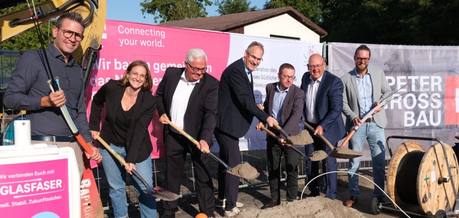 Beim Spatenstich in Herxheim (von links): Björn Klein (technische Projektleitung Telekom), SÜW-Gigabitbeauftragte Karolin Söltner, Gerd Schäfer (Regionalmanager Telekom), Landrat Dietmar Seefeldt, Thomas Kupfer (Fördermittelberater Aconium GmbH), Uwe König (Geschäftsführer der Mittelstands- und Betreuungsgesellschaft Südliche Weinstraße mbH) und Christian Roggenbuck (Gigabit-Kompetenzzentrum des Landes). Foto: KV SÜW Mehrere Personen mit Schaufeln führen einen symbolischen Spatenstich für den Glasfaserausbau durch. Im Hintergrund sind Banner mit Logos und Schriftzügen zu sehen, im Vordergrund liegt ein Sandhaufen.