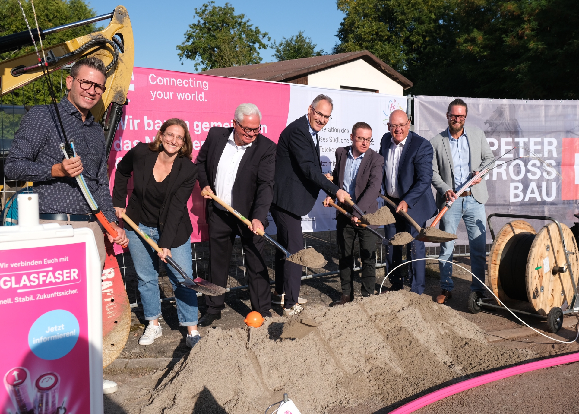 Mehrere Personen mit Schaufeln führen einen symbolischen Spatenstich für den Glasfaserausbau durch. Im Hintergrund sind Banner mit Logos und Schriftzügen zu sehen, im Vordergrund liegt ein Sandhaufen.