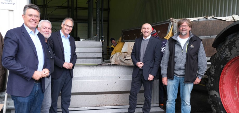 Fünf Männer stehen vor einer großen Edelstahlanlage in einer Halle. Neben ihnen ist ein Traktor mit Anhänger zu sehen. Die Gruppe blickt in die Kamera und lächelt.