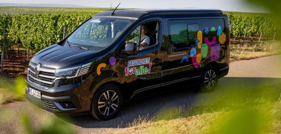 Ein schwarzer Kleinbus mit der Aufschrift "Gesundheit für alle" steht auf einem schmalen Weg vor einer Weinberglandschaft, die sich im Hintergrund bis zum Horizont erstreckt. Auch im Vordergrund erscheinen am Bildrand unscharf ein paar Blätter, sodass der Eindruck entsteht, es wurde durch die Reben fotografiert.