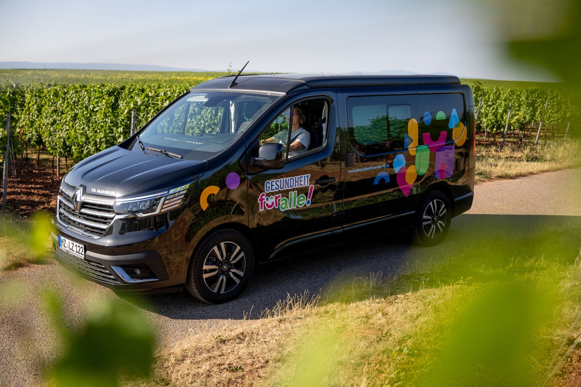 Ein schwarzer Kleinbus mit der Aufschrift "Gesundheit für alle" steht auf einem schmalen Weg vor einer Weinberglandschaft, die sich im Hintergrund bis zum Horizont erstreckt. Auch im Vordergrund erscheinen am Bildrand unscharf ein paar Blätter, sodass der Eindruck entsteht, es wurde durch die Reben fotografiert.