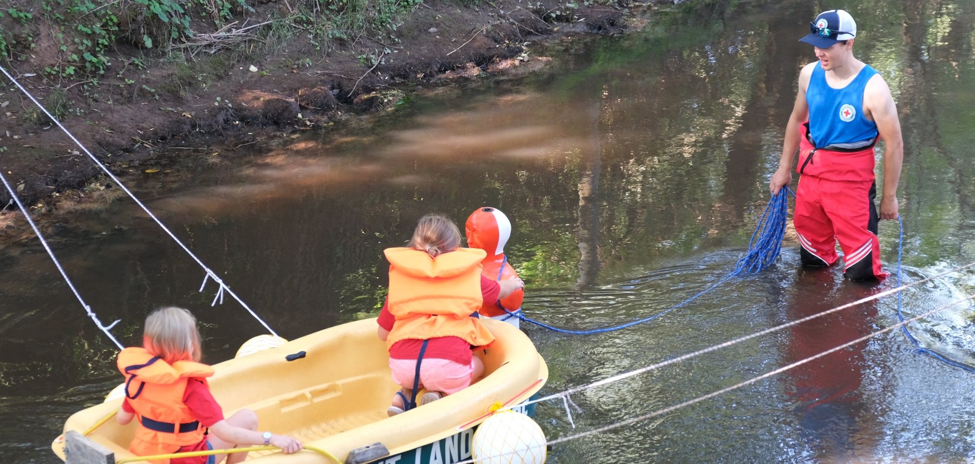 Zwei Kinder mit Schwimmwesten sitzen in einem kleinen Boot, das mit Seilen gesichert über einen Bach gezogen wird. Neben dem Boot steht ein Helfer im Wasser, der die Übung begleitet.