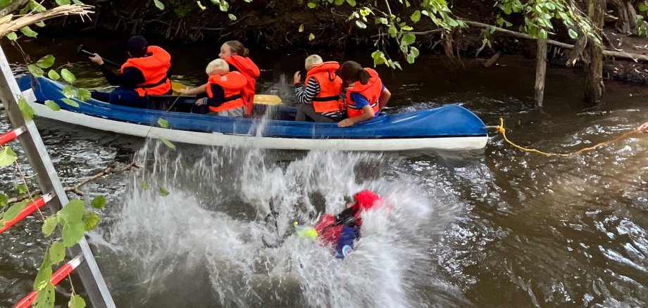 Platsch! Ein Mitglied der DRK Wasserwacht SÜW machte sich einen Spaß daraus, die Kanuten mit einem beherzten Sprung in die Queich nass zu spritzen. Foto: KV SÜW Fünf Kinder und Jugendliche fahren in einem Kanu, während ein Mitglied der DRK Wasserwacht SÜW sich einen Spaß daraus macht, die Kanuten mit einem beherzten Sprung in die Queich nass zu spritzen.