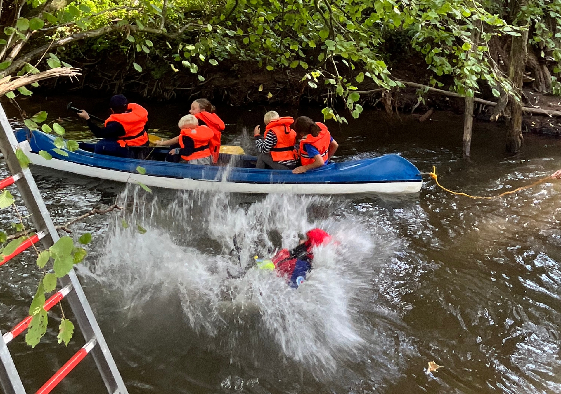 Fünf Kinder und Jugendliche fahren in einem Kanu, während ein Mitglied der DRK Wasserwacht SÜW sich einen Spaß daraus macht, die Kanuten mit einem beherzten Sprung in die Queich nass zu spritzen.