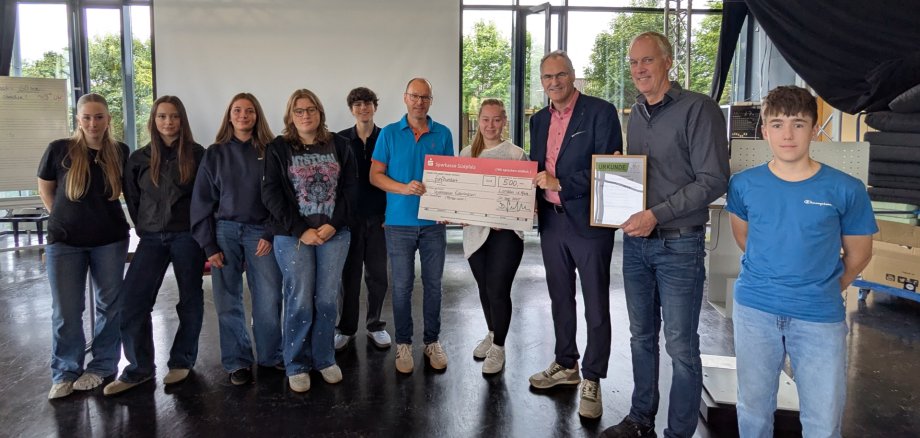 Gruppenbild mehrerer Erwachsener und Schüler. Zwei Männer und eine Frau halten einen großen symbolischen Scheck. Ein weiterer Mann hält eine Urkunde, die das Gymnasium Edenkoben als Gewinner des Schulradelns 2025 im Kreis SÜW auszeichnet.