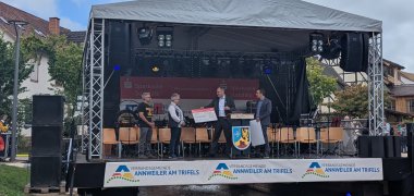 Zwei auf einen Streich: In Annweiler überreichte Landrat Dietmar Seefeldt (Zweiter von rechts) mit Verbandsgemeinde-Bürgermeister Christian Burkhart (rechts) einen Scheck an die Teamkapitänin beziehungsweise den -kapitän des Evangelischen Trifels-Gymnasiums und des Staufer-Schulzentrums. Foto: KV SÜW Auf einer Bühne, die über einen kleinen Flusslauf gebaut ist, überreichen zwei Männer je einen großen symbolischen Scheck an eine Frau und einen Mann.