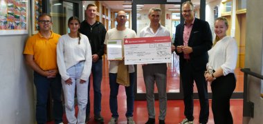 Landrat Dietmar Seefeldt (Zweiter von rechts) und SÜW-Klimaschutzmanagerin Agnes Bartmus (rechts) übergeben der Gebrüder-Ullrich-Realschule plus Maikammer-Hambach den Scheck fürs Schulradeln. Foto: Realschule plus Maikammer-Hambach Gruppenbild mehrerer Erwachsener und Schüler. Ein Mann hält einen großen symbolischen Scheck. Ein weiterer Mann hält eine Urkunde, die ddie Gebrüder-Ullrich-Realschule plus Maikammer-Hambach als erfolgreichen Teilnehmer des Schulradelns 2025 im Kreis SÜW auszeichnet.