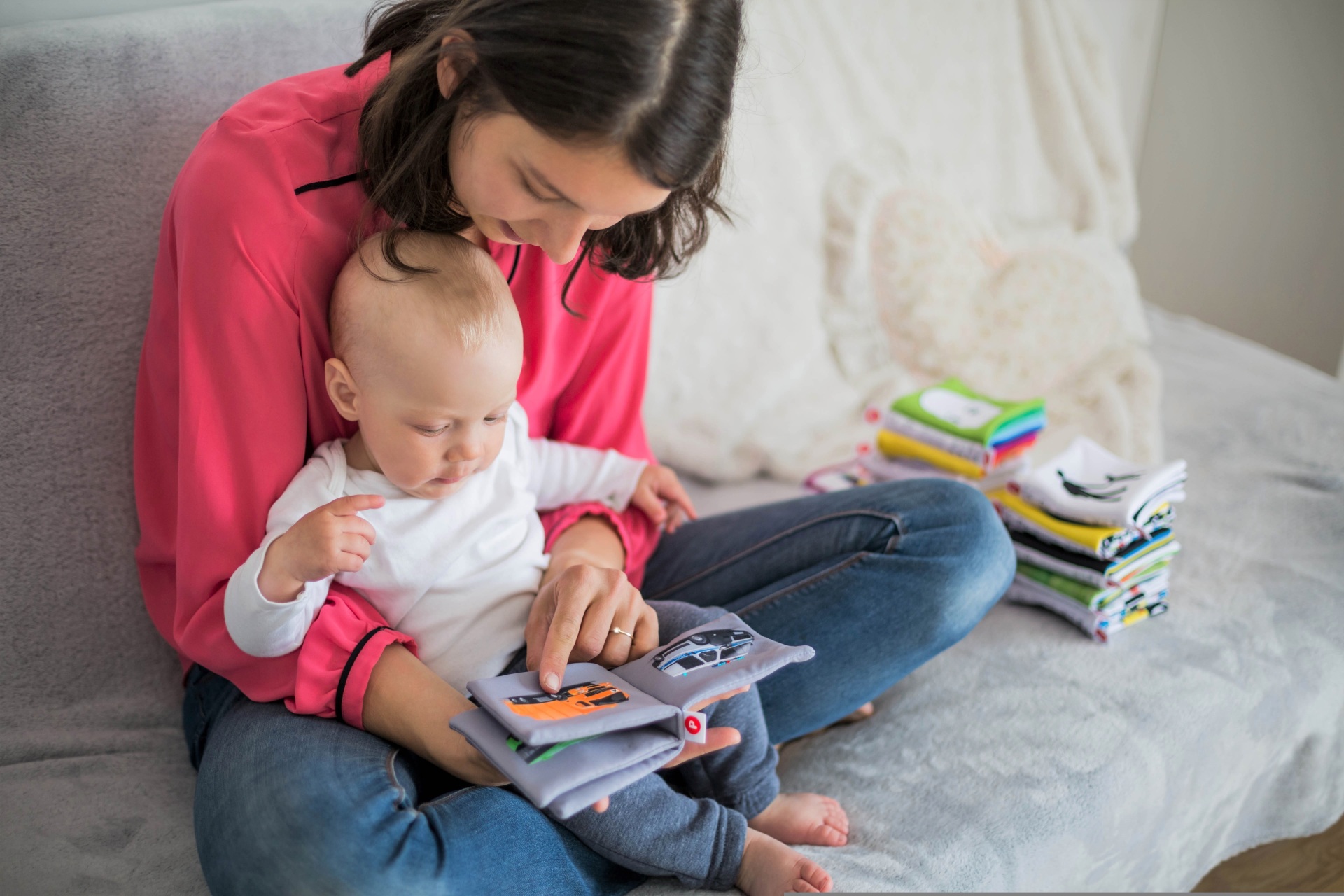 Eine Frau sitzt auf einem Sofa und schaut gemeinsam mit einem Baby ein kleines Bilderbuch an, während neben ihnen weitere Kinderbücher liegen.
