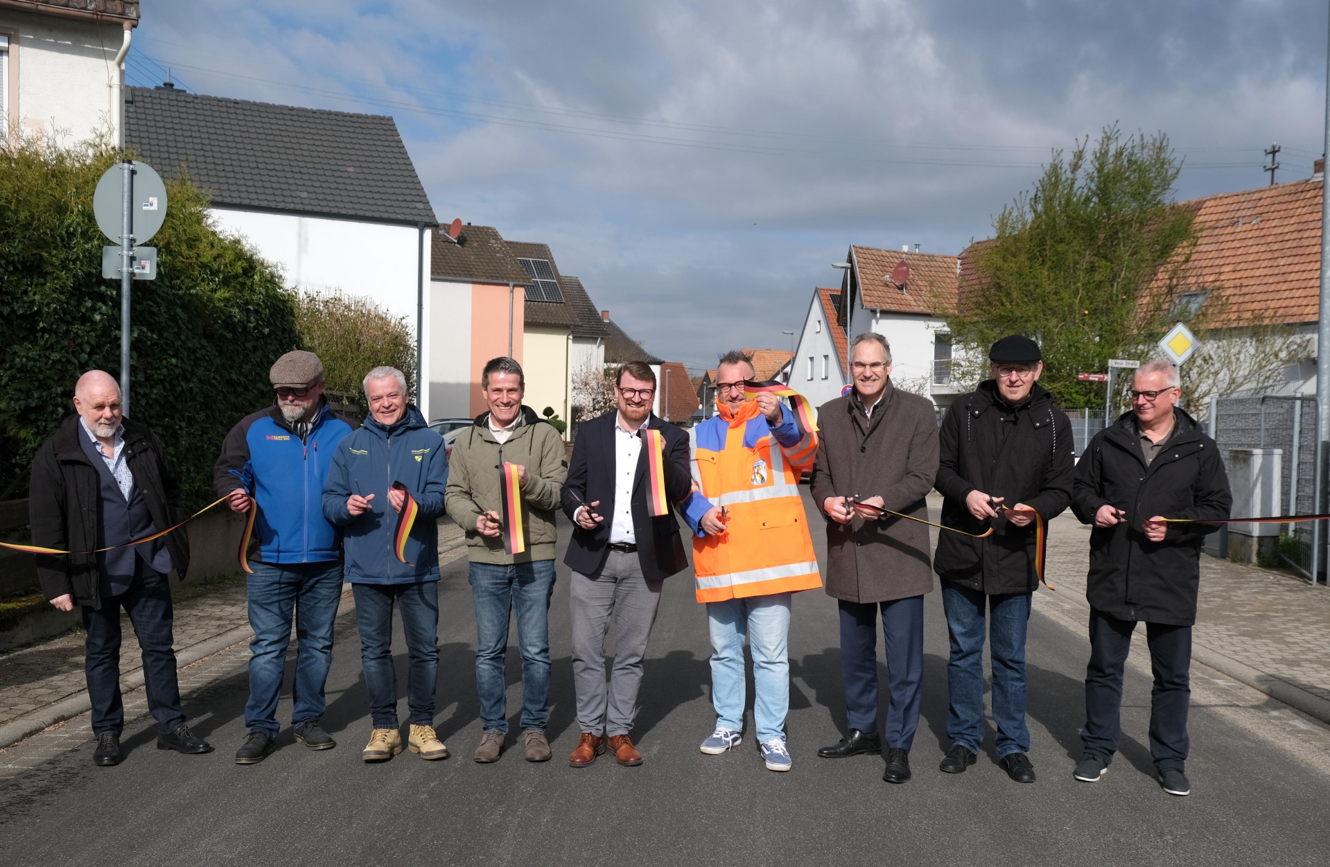 Gruppenbild: Neun Männer stehen innerorts nebeneinander auf einer Straße. Einige halten Scheren und Teile eines Bandes in den Farben der Deutschen Flagge, das sie zuvor durchschnitten haben. Damit geben sie symbolisch die Straße frei.