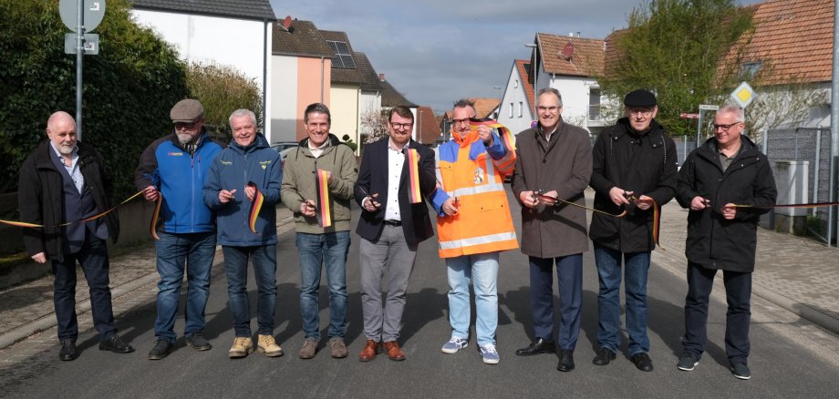 Gruppenbild: Neun Männer stehen innerorts nebeneinander auf einer Straße. Einige halten Scheren und Teile eines Bandes in den Farben der Deutschen Flagge, das sie zuvor durchschnitten haben. Damit geben sie symbolisch die Straße frei.