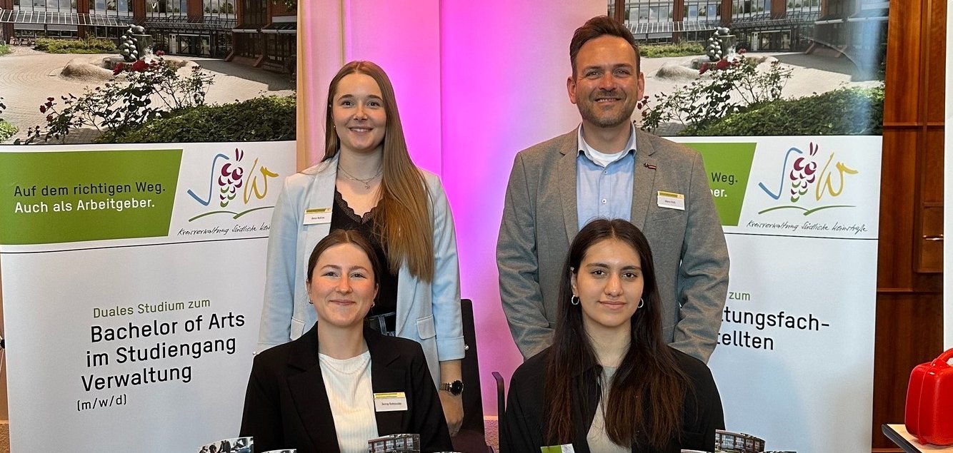 Vier Personen aus dem Team der Kreisverwaltung SÜW an einem Messestand. Auf einem Tisch liegen Info-Flyer. Im Hintergrund sind zwei Rollups zu sehen, die das duale Studium und die Ausbildung bei der Kreisverwaltung bewerben.