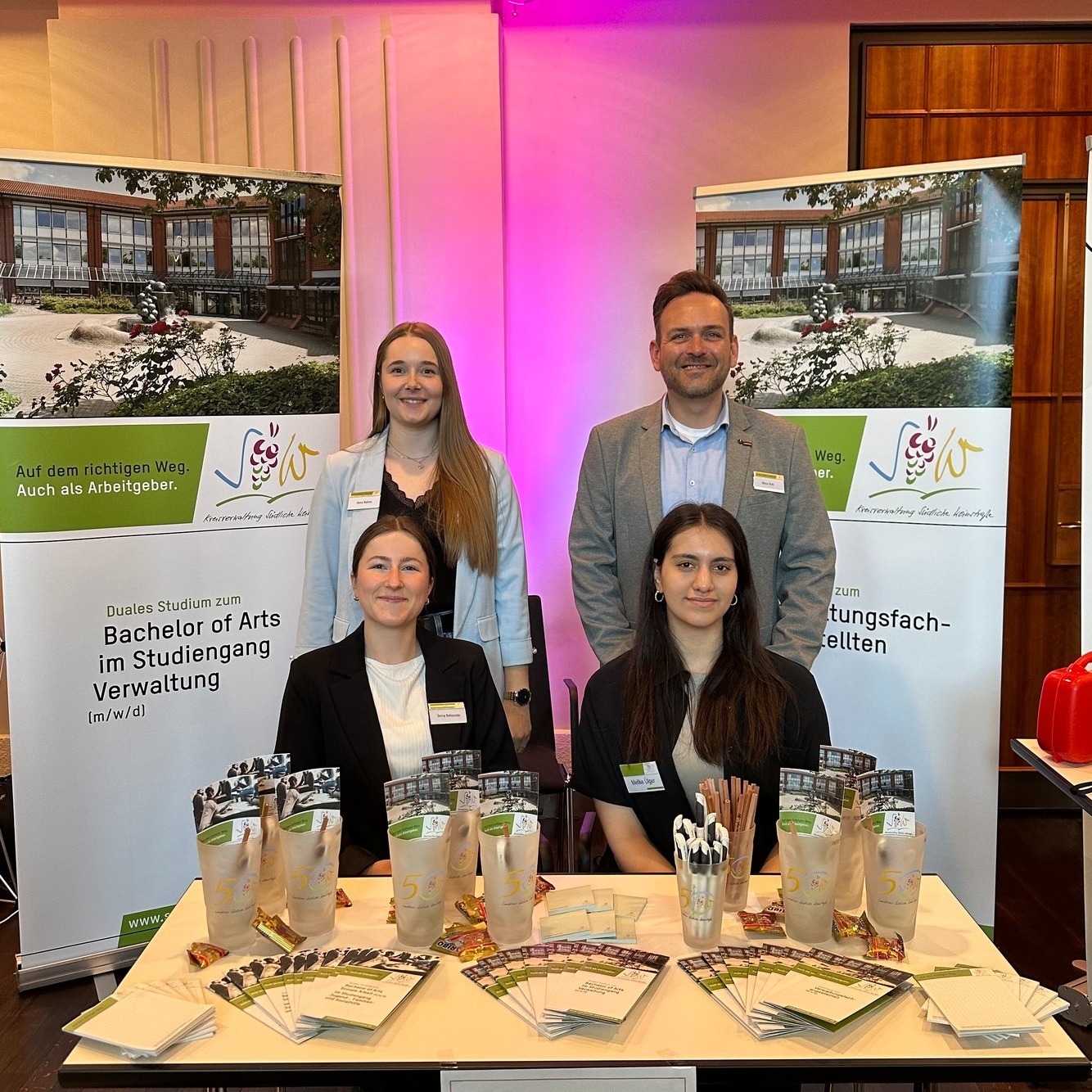 Vier Personen aus dem Team der Kreisverwaltung SÜW an einem Messestand. Auf einem Tisch liegen Info-Flyer. Im Hintergrund sind zwei Rollups zu sehen, die das duale Studium und die Ausbildung bei der Kreisverwaltung bewerben.