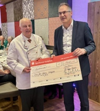 Zwei Männer, einer in weißer Schürze , einer im Anzug, halten einen übergroßen, symbolischen Spendenscheck.