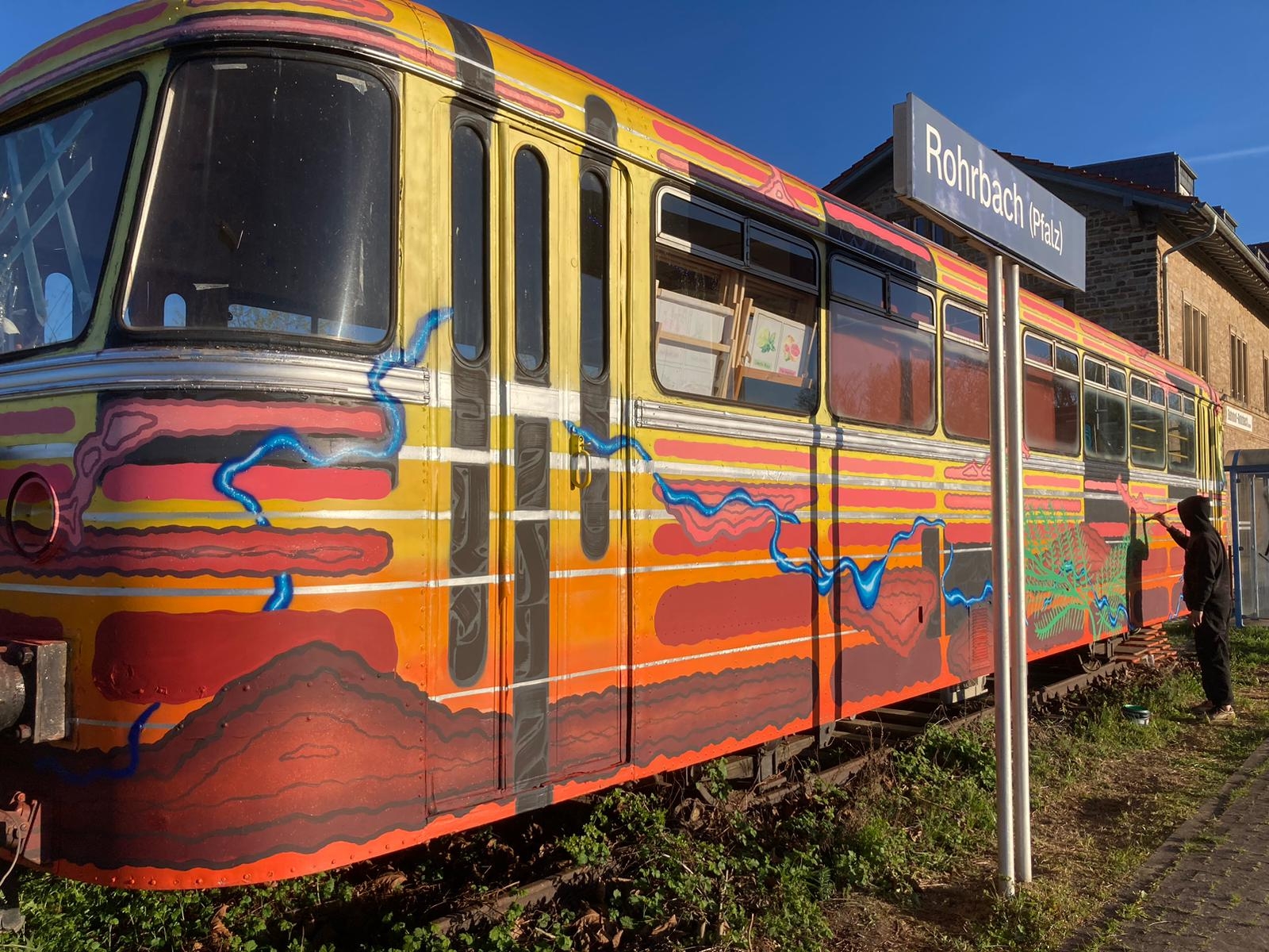 Ein bunter Eisenbahnwagen, eine Person mit schwarzer Kapuze, Bahnhofsschild mit Aufschrift "Rohrbach".