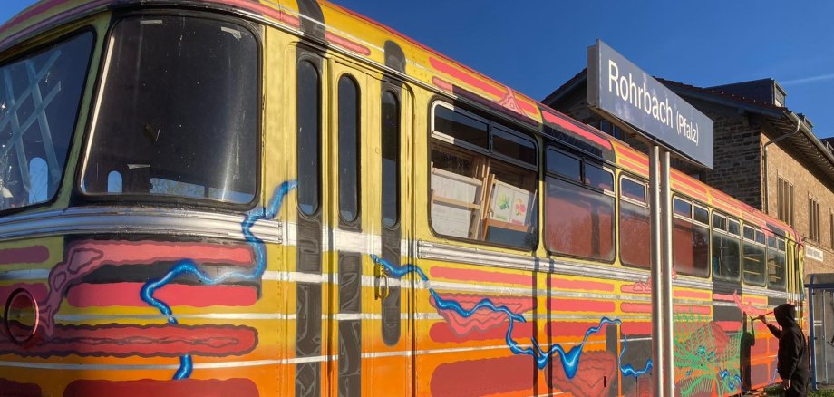 Der alte Triebwagen am Bahnhof Rohrbach wird derzeit von einer Künstlergruppe umgestaltet. Foto: Karlheinz Zwick Ein bunter Eisenbahnwagen, eine Person mit schwarzer Kapuze, Bahnhofsschild mit Aufschrift "Rohrbach".