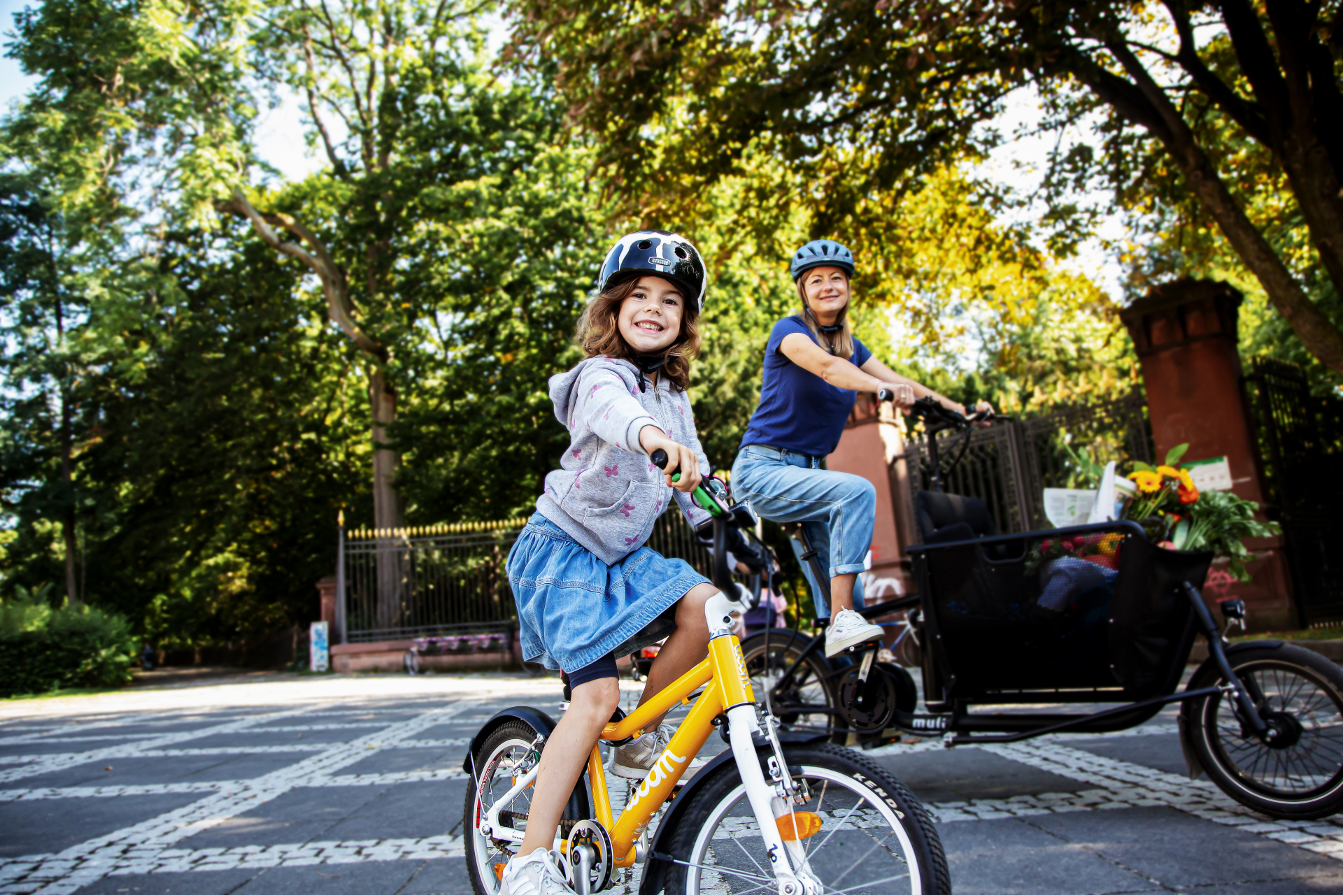 Ein Mädchen fährt mit Helm auf einem gelben Fahrrad über eine Straße und lächelt in die Kamera. Hinter ihr fährt eine Frau mit Helm auf einem Lastenrad durch eine grüne, von Bäumen gesäumte Straße.