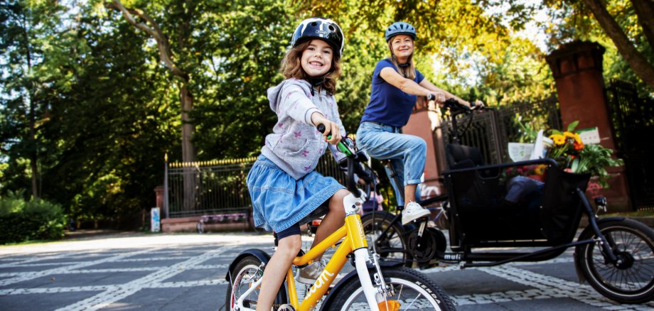 Ein Mädchen fährt mit Helm auf einem gelben Fahrrad über eine Straße und lächelt in die Kamera. Hinter ihr fährt eine Frau mit Helm auf einem Lastenrad durch eine grüne, von Bäumen gesäumte Straße.