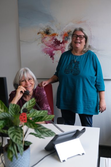 Zwei ältere Frauen in einem Büro: Eine sitzt am Schreibtisch und telefoniert lächelnd, die andere steht daneben und lacht. Im Vordergrund steht eine Zimmerpflanze mit Marienkäfer-Deko, im Hintergrund hängt ein abstraktes, farbenfrohes Gemälde.