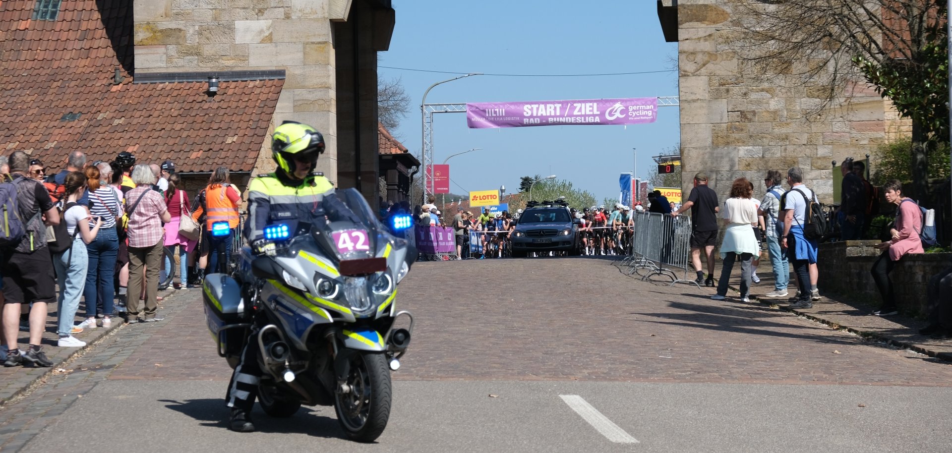 Ein Polizeimotorrad fährt vor dem Deutschen Weintor in Schweigen-Rechtenbach, während dahinter ein Radrennen startet. Unter einem Banner mit der Aufschrift "Start/ziel Rad-Bundesliga" warten zahlreiche Radfahrerinnen und Radfahrer am Start. Zuschauerinnen und Zuschauer stehen beidseitig der Straße hinter Absperrungen und verfolgen das Geschehen.