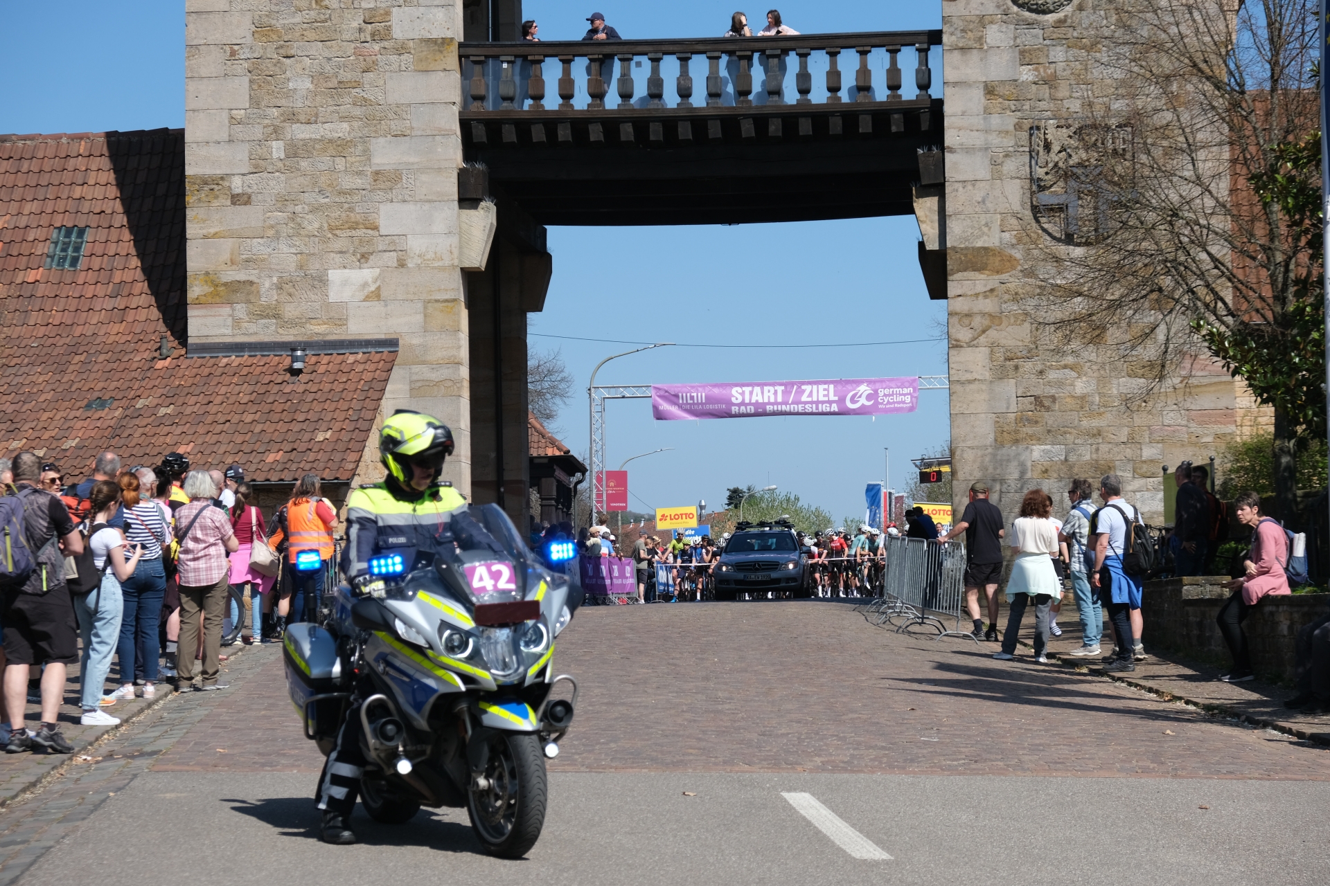 Ein Polizeimotorrad fährt vor dem Deutschen Weintor in Schweigen-Rechtenbach, während dahinter ein Radrennen startet. Unter einem Banner mit der Aufschrift "Start/ziel Rad-Bundesliga" warten zahlreiche Radfahrerinnen und Radfahrer am Start. Zuschauerinnen und Zuschauer stehen beidseitig der Straße hinter Absperrungen und verfolgen das Geschehen.
