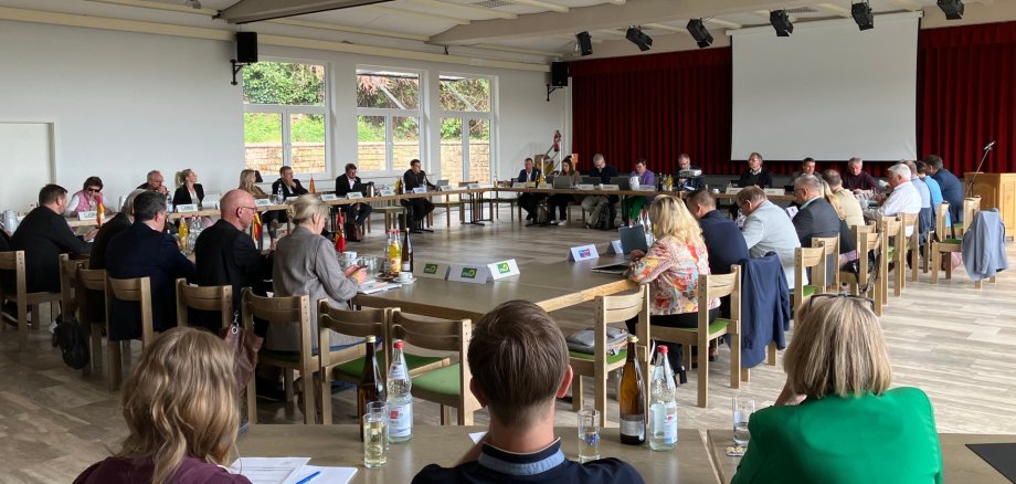 Blick in den Kreistag. Die versammelten Mitglieder sitzen in einem hellen Saal an im Quadrat angeordneten Tischen.