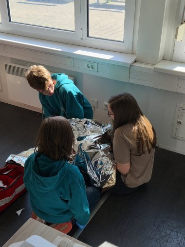 Drei Jugendliche knien in einem hellen Raum am Boden und versorgen eine Person, die mit einer Rettungsdecke abgedeckt ist.