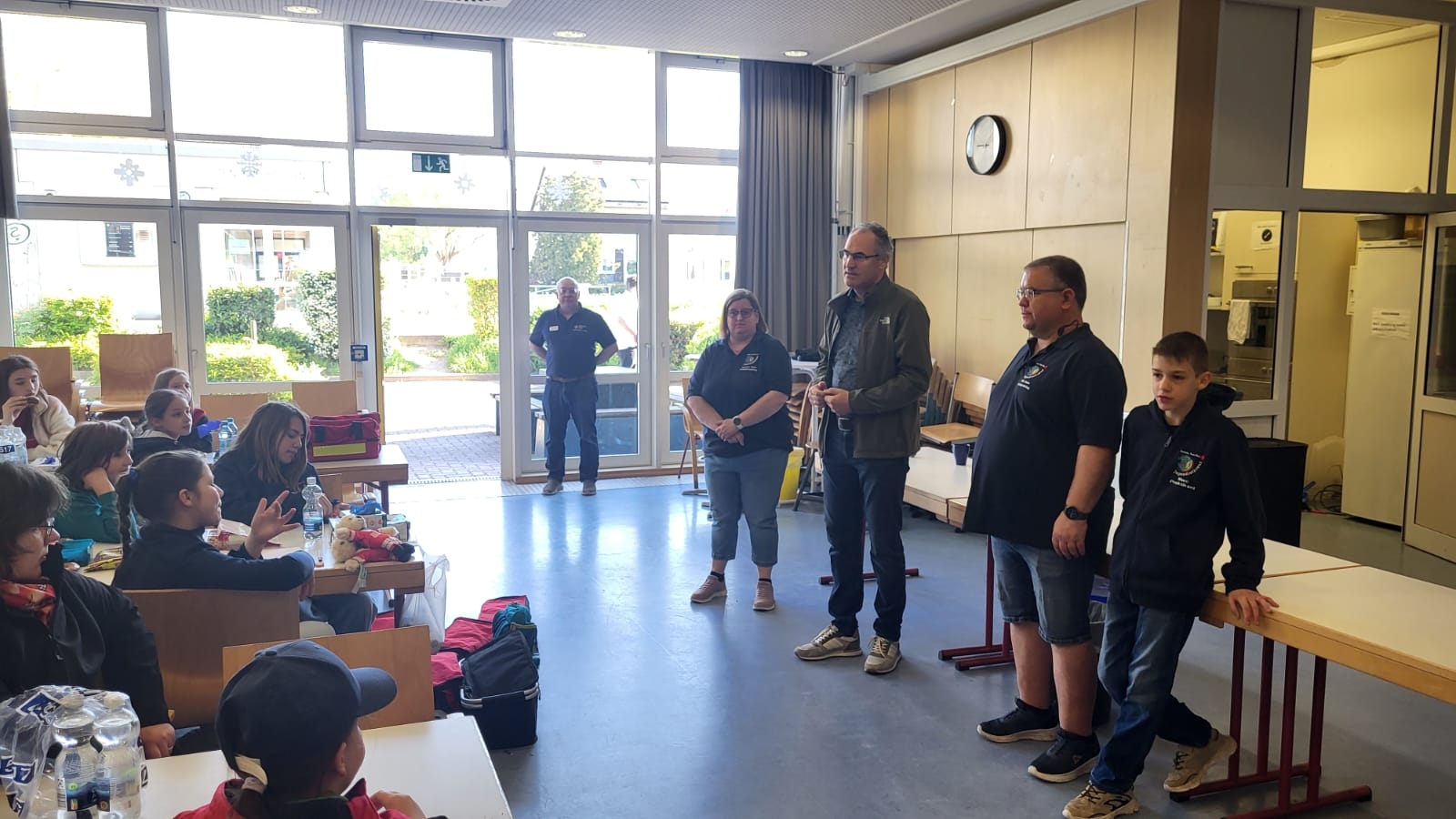 Mehrere Kinder und Jugendliche sitzen in einem Raum an Tischen, während vorne Erwachsene und ein Jugendlicher stehen und offenbar eine Gruppe ansprechen.