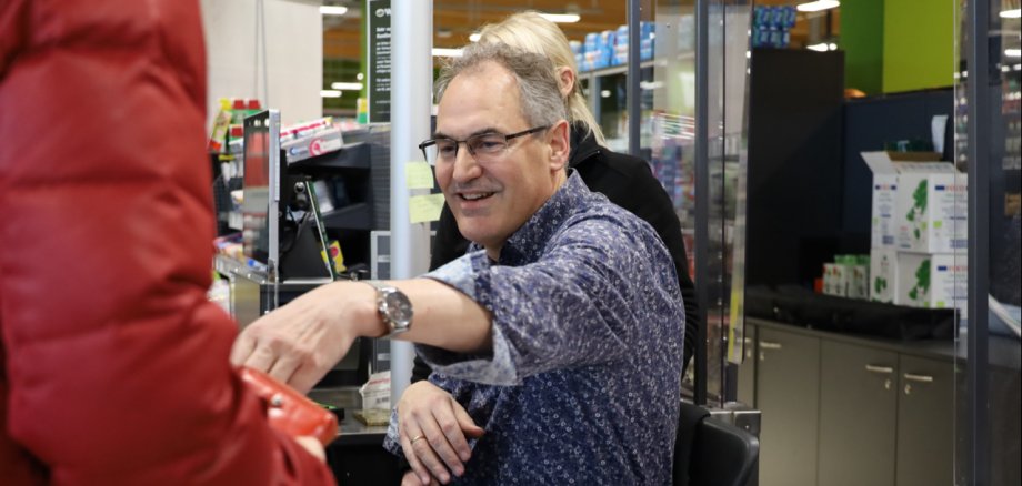 Landrat Dietmar Seefeldt sitzt an einer Supermarktkasse und gibt Wechselgeld an eine Person in roter Jacke, deren rechter Arm im Vordergrund des Bildes zu erkennen ist.