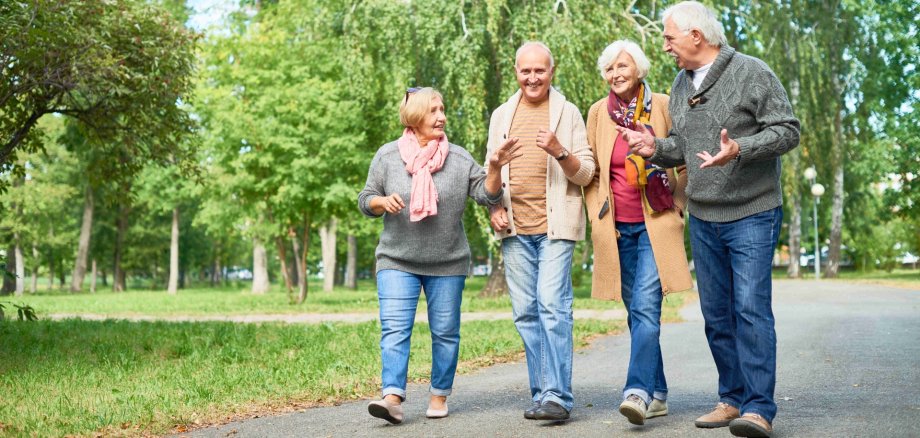 Eine Gruppe Seniorinnen und Senioren in warmer Kleidung läuft während einer freudigen Unterhaltung durch einen Park.