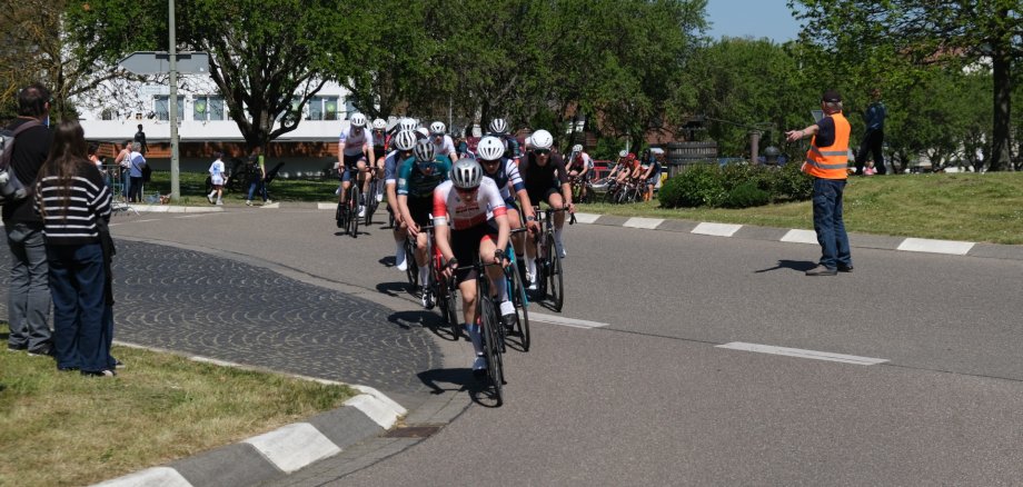 Radfahrer fahren aus einem Kreisverkehr aus auf die Zielgerade des Rennens. Ein Ordner in orangener Warnweste zeigt ihnen die zu nehmende Ausfahrt an.