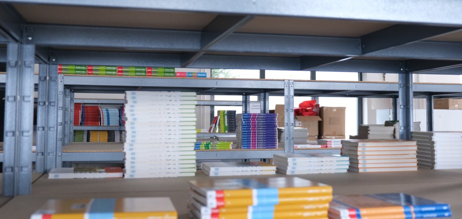 Gestapelte Schulbücher liegen auf einem Regal in einem Lagerraum. Im Hintergrund sind weitere Bücherregale mit Schulbüchern zu sehen.