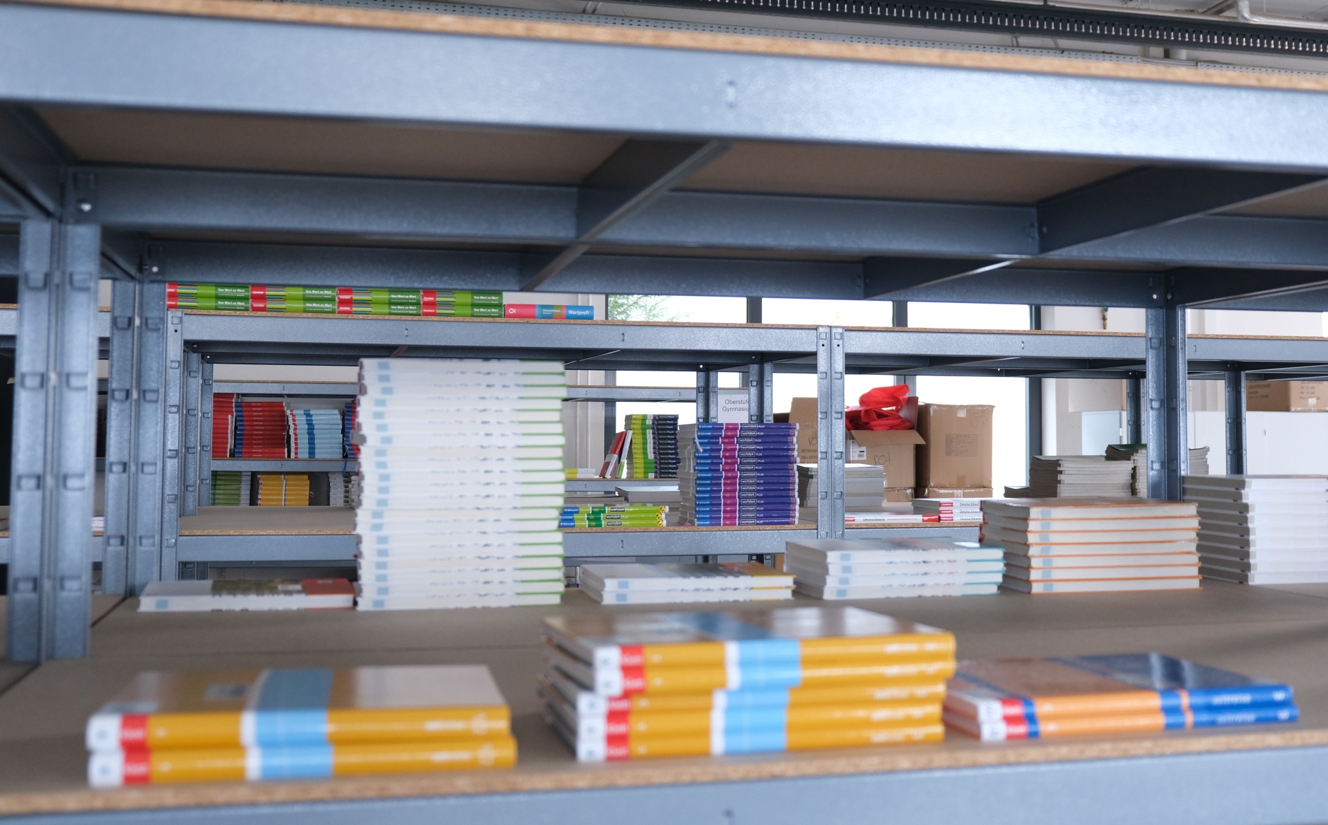 Gestapelte Schulbücher liegen auf einem Regal in einem Lagerraum. Im Hintergrund sind weitere Bücherregale mit Schulbüchern zu sehen.