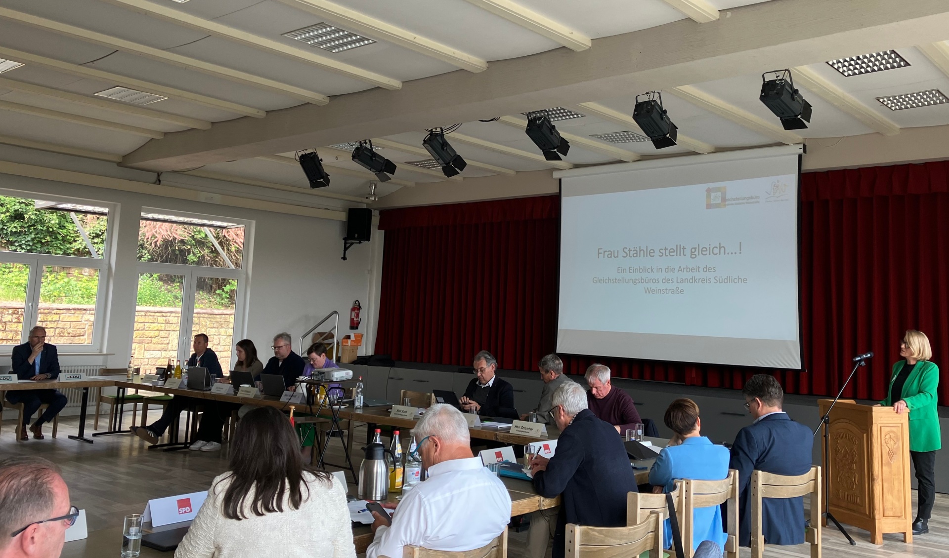 Kreistagsmitglieder sitzen an im Quadrat angeordneten Tischen in einem hellen Saal und hören den von der Gleichstellungsbeauftragen Isabelle Stähle vorgetragenen Bericht. Die Rednerin steht rechts im Bild an einem Rednerpult.