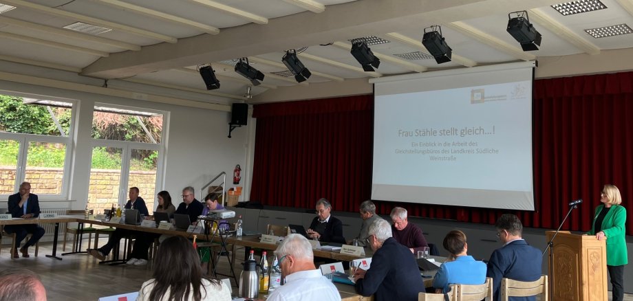 Kreistagsmitglieder sitzen an im Quadrat angeordneten Tischen in einem hellen Saal und hören den von der Gleichstellungsbeauftragen Isabelle Stähle vorgetragenen Bericht. Die Rednerin steht rechts im Bild an einem Rednerpult.