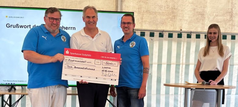 Spendenübergabe an den Verein Team Bananenflanke e.V. Foto: Seefeldt Drei Männer stehen auf einer Bühne und präsentieren gemeinsam einen Spendenscheck über 500 Euro für das Team Bananenflanke. Rechts daneben steht eine Frau an einem Stehtisch mit Mikrofon.