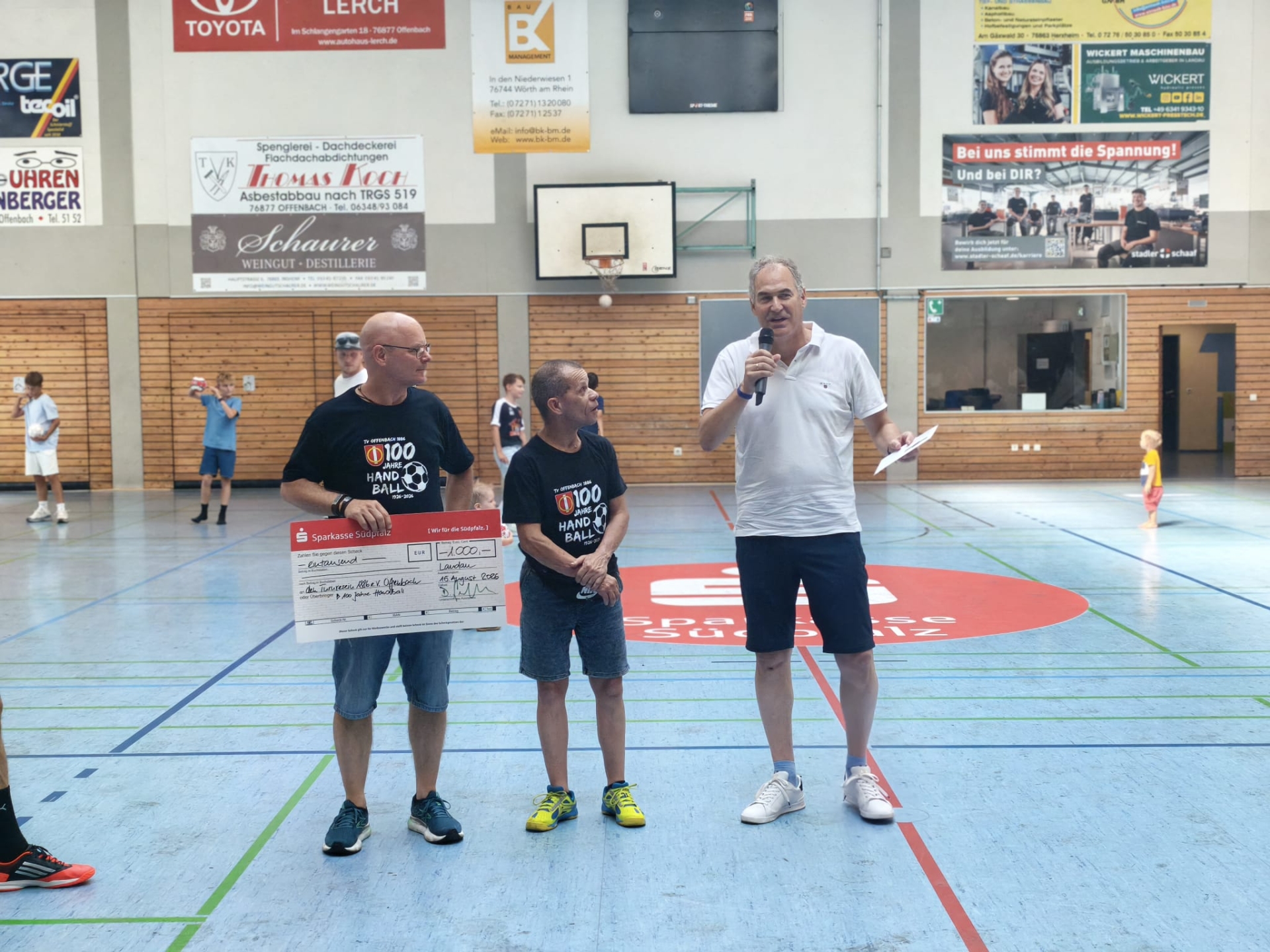 Drei Männer stehen in einer Sporthallte. Einer hält einen Spendenscheck über 1000 Euro, währen ein weiterer Mann mit einem Mikrofon zum Publikum spricht. Im Hintergrund sind weitere Personen auf dem Spielfeld zu sehen.