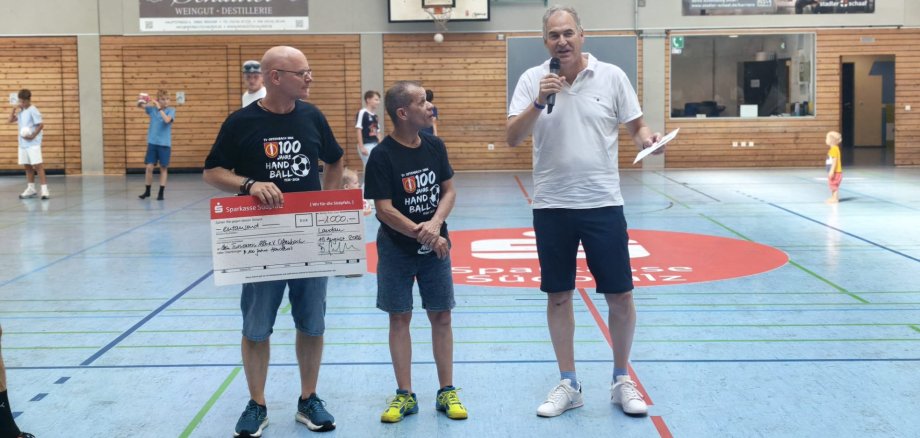 Spendenübergabe an die Abteilung Handball des Turnverein 1886 e.V. Offenbach. Foto: Seefeldt Drei Männer stehen in einer Sporthallte. Einer hält einen Spendenscheck über 1000 Euro, währen ein weiterer Mann mit einem Mikrofon zum Publikum spricht. Im Hintergrund sind weitere Personen auf dem Spielfeld zu sehen.