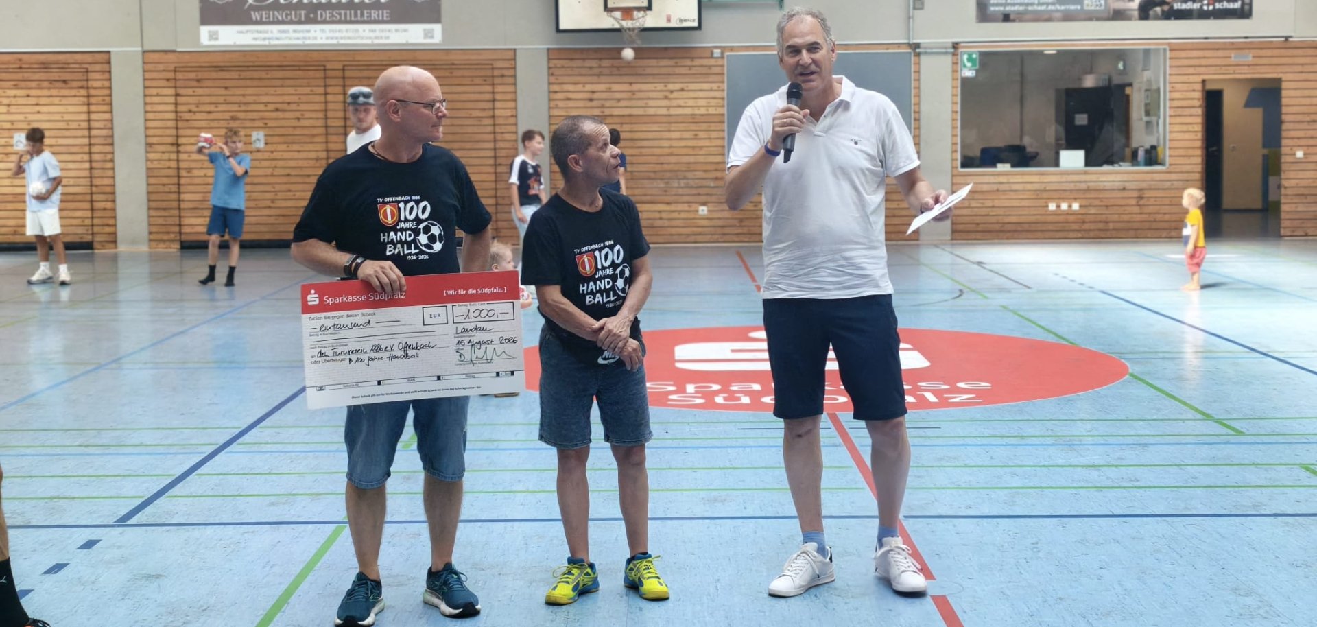 Drei Männer stehen in einer Sporthallte. Einer hält einen Spendenscheck über 1000 Euro, währen ein weiterer Mann mit einem Mikrofon zum Publikum spricht. Im Hintergrund sind weitere Personen auf dem Spielfeld zu sehen.