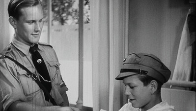 Screenshot aus einem Schwarz-Weiß-Film: Ein uniformierter Mann steht neben einem Jungen mit Schirmmütze. Beide blicken nach unten auf etwas außerhalb des Bildausschnitts.