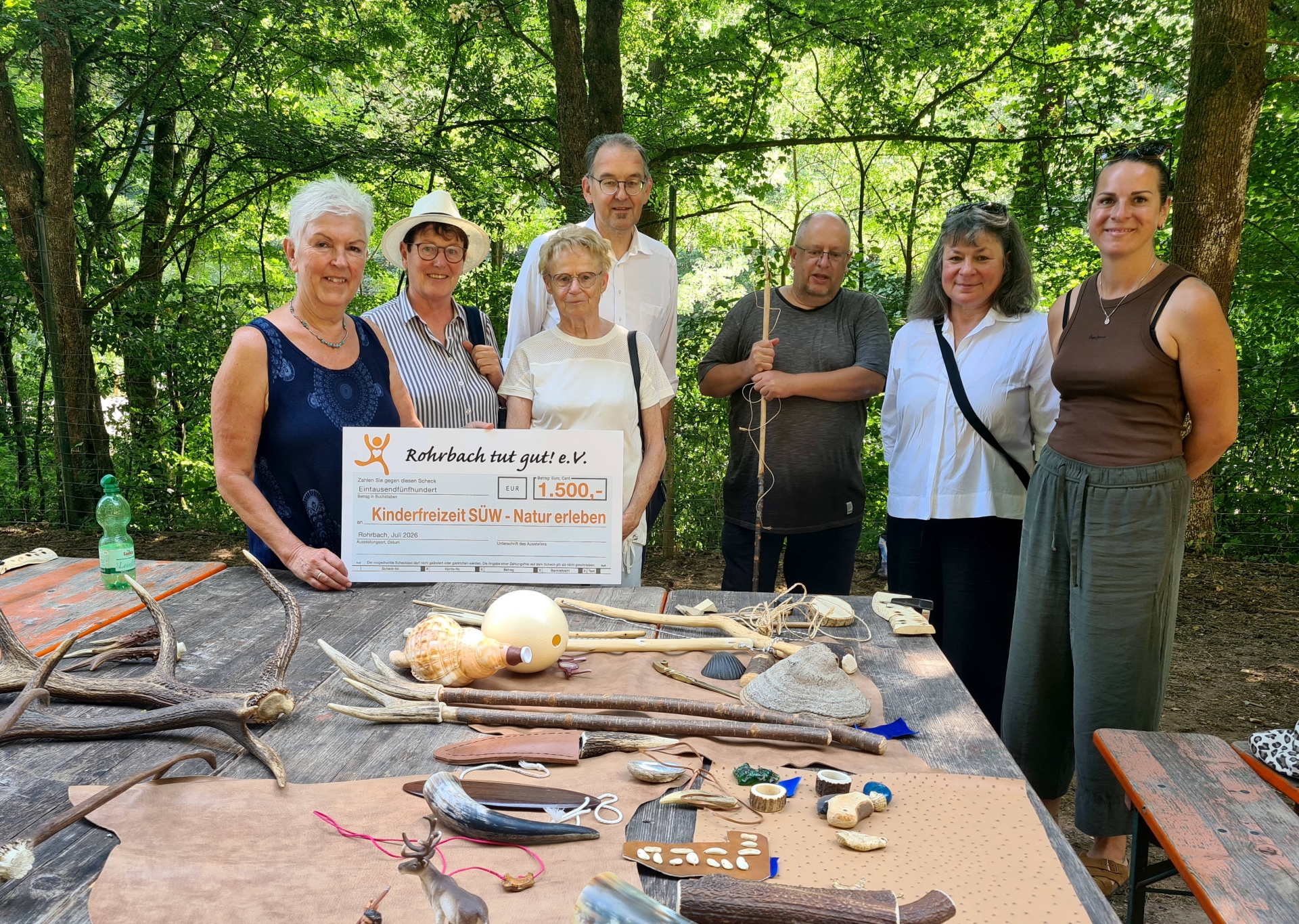 Sieben Personen stehen im Freien an einem Tisch in einem bewaldeten Gelände. Zwei Frauen halten einen symbolischen Spendenscheck.