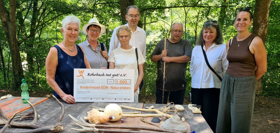 Von links nach rechts: Andrea Weng, Tilla Schneider und Christel Gaschler von Rohrbach tut gut! e.V. mit Erstem Kreisbeigeordneten Georg Kern, Freizeitleiter Andreas Stern, Jugendamtsleiterin Hannelore Schlageter und Nina Keßler, Leiterin des Allgemeinen Sozialen Dienstes des Jugendamts. Foto: KV SÜW Sieben Personen stehen im Freien an einem Tisch in einem bewaldeten Gelände. Zwei Frauen halten einen symbolischen Spendenscheck.